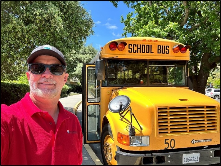 A smiling man standing outside next to a yellow school bus.