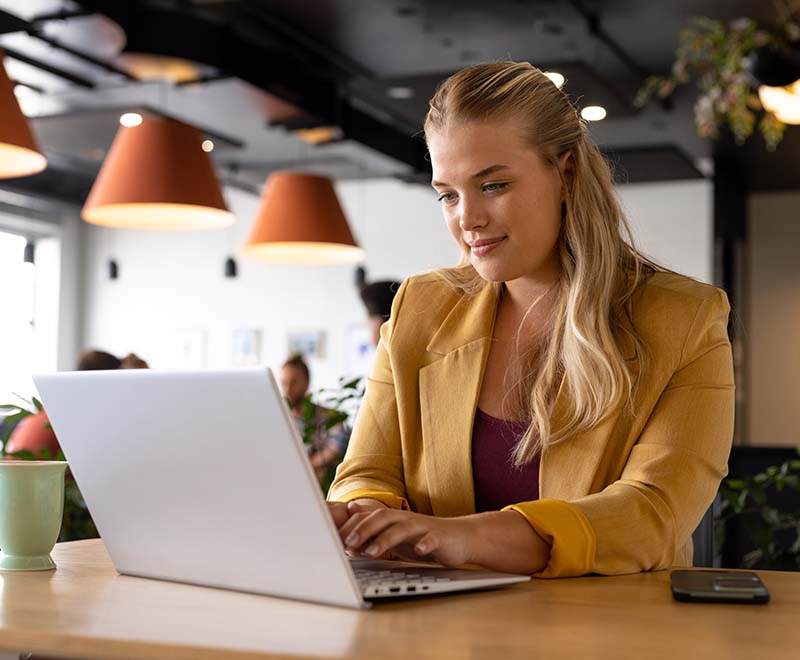 A woman sits in an open workspace, focused on working on her laptop.