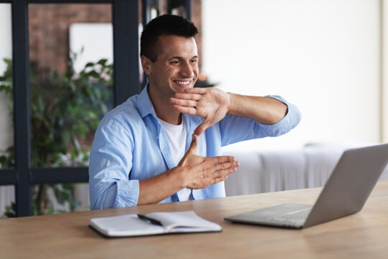 A man sitting at a desk and smiling while gesturing with his hands during a video call on his laptop.